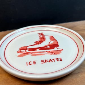 May include: A small, round, white ceramic dish with a red border. The dish features a red illustration of an ice skate and the words "ICE SKATES" in red. The dish is on a wooden surface.