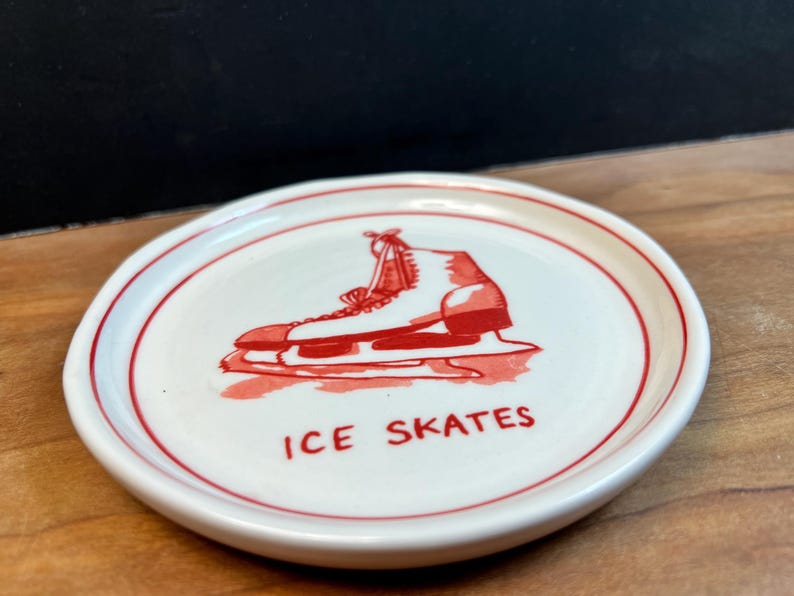 May include: A small, round, white ceramic dish with a red border. The dish features a red illustration of an ice skate and the words "ICE SKATES" in red. The dish is on a wooden surface.