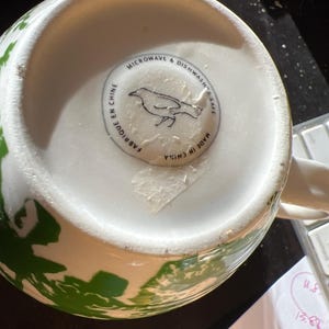 May include: White ceramic mug with green floral design. The bottom of the mug has a sticker with a bird illustration and text that reads "MICROWAVE & DISHWASHER SAFE", "FABRIQUE EN CHINE", and "MADE IN CHINA".
