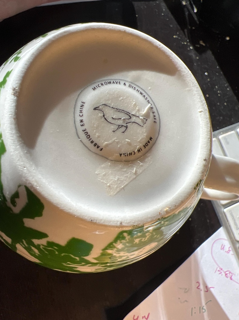 May include: White ceramic mug with green floral design. The bottom of the mug has a sticker with a bird illustration and text that reads "MICROWAVE & DISHWASHER SAFE", "FABRIQUE EN CHINE", and "MADE IN CHINA".