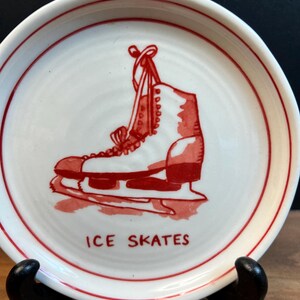 May include: A white ceramic plate with a red border and a red illustration of an ice skate. The words "ICE SKATES" are printed in red below the skate. The plate is displayed on a black stand.
