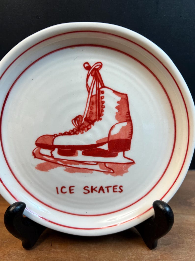 May include: A white ceramic plate with a red border and a red illustration of an ice skate. The words "ICE SKATES" are printed in red below the skate. The plate is displayed on a black stand.