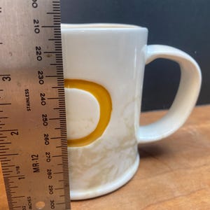 Puede incluir: Una taza de cer&aacute;mica blanca con la letra "D" amarilla en el lateral. La taza mide aproximadamente 7,6 cm de alto. La taza tiene un asa curva y un patr&oacute;n beige claro en la mitad inferior.