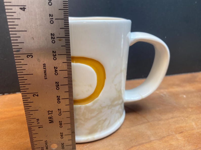 Puede incluir: Una taza de cer&aacute;mica blanca con la letra "D" amarilla en el lateral. La taza mide aproximadamente 7,6 cm de alto. La taza tiene un asa curva y un patr&oacute;n beige claro en la mitad inferior.