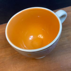 K&ouml;nnte beinhalten: Eine wei&szlig;e Keramik-Teetasse mit orangefarbenem Innenraum und kleinem Henkel. Der Rand ist mit einer d&uuml;nnen Goldlinie und einem zarten Blumenmuster verziert. Die Tasse steht auf einer Holzoberfl&auml;che.