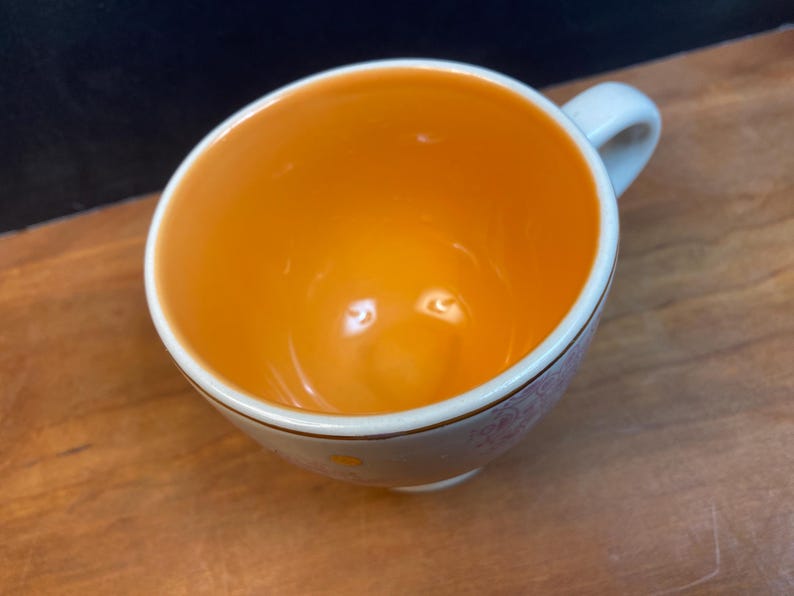 K&ouml;nnte beinhalten: Eine wei&szlig;e Keramik-Teetasse mit orangefarbenem Innenraum und kleinem Henkel. Der Rand ist mit einer d&uuml;nnen Goldlinie und einem zarten Blumenmuster verziert. Die Tasse steht auf einer Holzoberfl&auml;che.