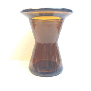 以下が含まれることがあります： 茶色のガラスの花瓶。砂時計型で、上部と底部が広く、中央が細くなっています。 花瓶は光沢のある仕上がりで、白い背景に置かれています。 ガラスは透明で、光を通します。