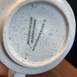May include: White ceramic mug with black speckles. The base of the mug features the text "by Anthropologie" and "Dishwasher & Microwave Safe Made in China / Fabriqu&eacute; en Chine."