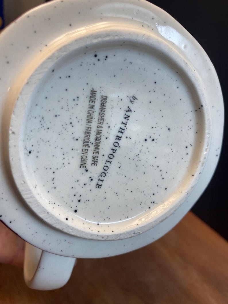 May include: White ceramic mug with black speckles. The base of the mug features the text "by Anthropologie" and "Dishwasher & Microwave Safe Made in China / Fabriqu&eacute; en Chine."