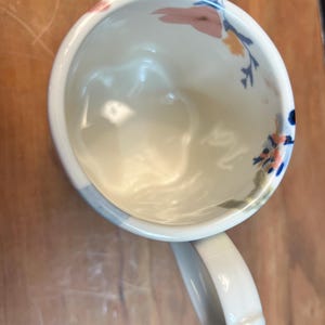 May include: White ceramic mug with a curved handle, decorated with floral patterns in pink, blue, and yellow. The mug's interior is white, and the handle has a rounded shape. The mug is set against a wooden surface.