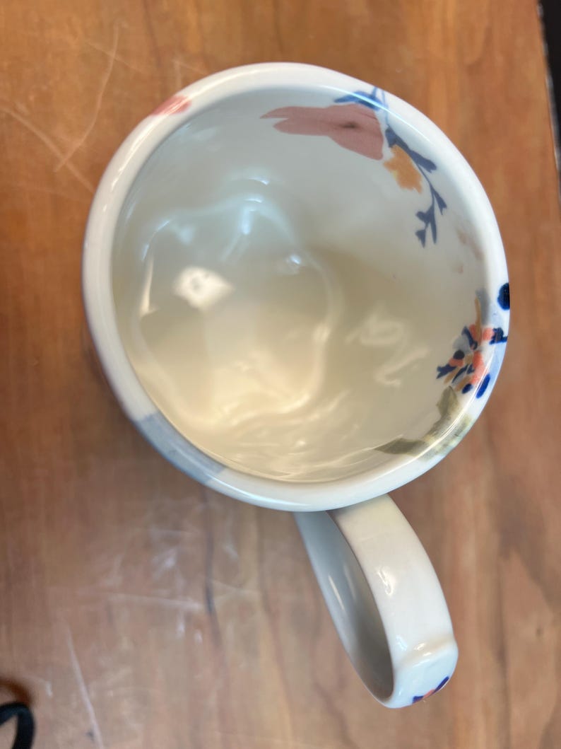 May include: White ceramic mug with a curved handle, decorated with floral patterns in pink, blue, and yellow. The mug's interior is white, and the handle has a rounded shape. The mug is set against a wooden surface.