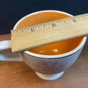 K&ouml;nnte beinhalten: Wei&szlig;e Teetasse mit orangefarbenem Innenraum und rosa Blumendesign. Ein Holzlineal misst etwa 5 cm &uuml;ber die Tasse. Die Tasse hat einen wei&szlig;en Henkel und einen goldenen Rand.