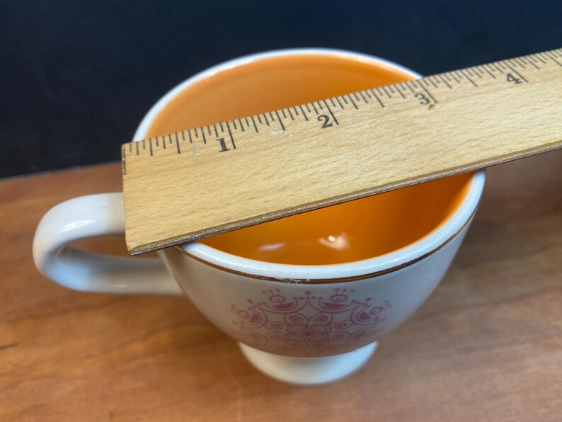 K&ouml;nnte beinhalten: Wei&szlig;e Teetasse mit orangefarbenem Innenraum und rosa Blumendesign. Ein Holzlineal misst etwa 5 cm &uuml;ber die Tasse. Die Tasse hat einen wei&szlig;en Henkel und einen goldenen Rand.