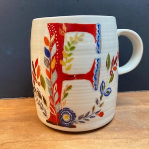 Può includere: Tazza in ceramica bianca con un motivo floreale rosso, blu e verde e la lettera "E" in rosso.