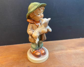 May include: A ceramic figurine of a child holding a lamb. The child wears a green hat, brown coat, orange scarf, green trousers, and orange shoes. The figurine is on a round, off-white base.