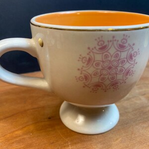K&ouml;nnte beinhalten: Cremefarbene Teetasse mit rosa Blumenmuster und Goldrand. Der Innenraum ist orange. Die Tasse hat einen Sockel und einen gebogenen Henkel. Ein kleiner Goldakzent befindet sich an der Seite der Tasse.