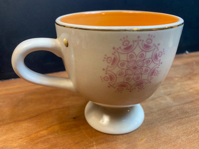 K&ouml;nnte beinhalten: Cremefarbene Teetasse mit rosa Blumenmuster und Goldrand. Der Innenraum ist orange. Die Tasse hat einen Sockel und einen gebogenen Henkel. Ein kleiner Goldakzent befindet sich an der Seite der Tasse.