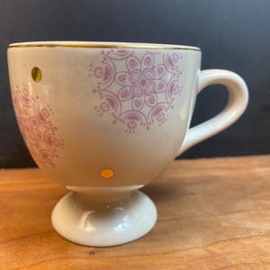 K&ouml;nnte beinhalten: Cremefarbene Teetasse mit Sockel, verziert mit rosa Blumenmustern und goldenen Akzenten. Die Tasse hat einen Henkel und zwei kleine goldene Punkte. Der Rand ist mit Gold verziert. Ein dekoratives St&uuml;ck zum Servieren von Tee oder Kaffee.