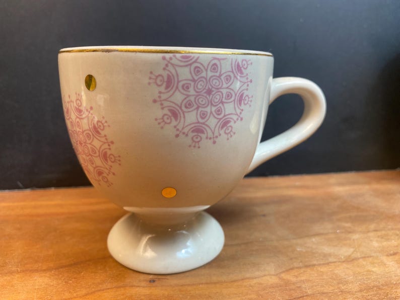 K&ouml;nnte beinhalten: Cremefarbene Teetasse mit Sockel, verziert mit rosa Blumenmustern und goldenen Akzenten. Die Tasse hat einen Henkel und zwei kleine goldene Punkte. Der Rand ist mit Gold verziert. Ein dekoratives St&uuml;ck zum Servieren von Tee oder Kaffee.