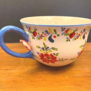 K&ouml;nnte beinhalten: Wei&szlig;e Keramik-Teetasse mit blauem Henkel und Rand, verziert mit einem Blumenmuster in Rot, Gelb und Blau. Der Henkel hat einen kleinen dekorativen Akzent. Die Tasse steht auf einer Holzoberfl&auml;che.