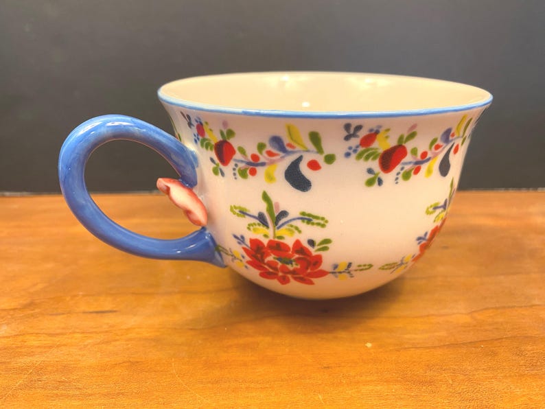 K&ouml;nnte beinhalten: Wei&szlig;e Keramik-Teetasse mit blauem Henkel und Rand, verziert mit einem Blumenmuster in Rot, Gelb und Blau. Der Henkel hat einen kleinen dekorativen Akzent. Die Tasse steht auf einer Holzoberfl&auml;che.