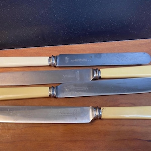 May include: Four vintage butter knives with cream-colored handles and silver blades. The knives are arranged on a wooden surface. Each knife has a silver bolster where the handle meets the blade. The blades have a slightly curved shape.