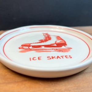 May include: A small, round, white ceramic dish with a red border and a red illustration of an ice skate. The words "ICE SKATES" are printed in red below the image. The dish is likely a decorative item or a small serving piece.