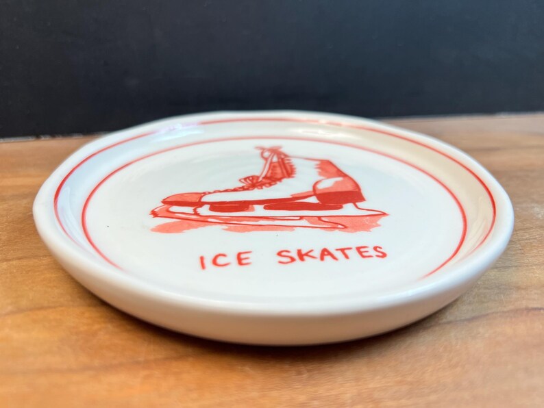 May include: A small, round, white ceramic dish with a red border and a red illustration of an ice skate. The words "ICE SKATES" are printed in red below the image. The dish is likely a decorative item or a small serving piece.