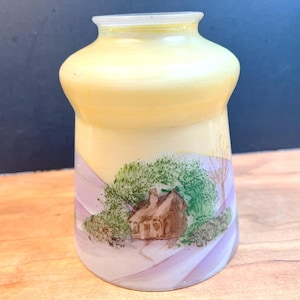 Może przedstawiać: Żółto-biały szklany wazon z namalowanym krajobrazem. Scena przedstawia brązową chatę pośród zielonych drzew i fioletowych wzgórz. Wazon ma zaokrąglony korpus i wąską szyję.