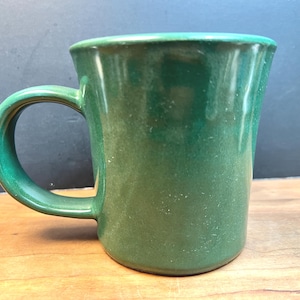Puede incluir: Una taza de cerámica verde lisa con un asa curva. La taza tiene un borde ligeramente ensanchado y un acabado liso y brillante. La taza está sobre una superficie de madera.