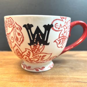 May include: White ceramic mug with a red handle and floral design. The mug features a black letter "W" and red floral patterns. The mug sits on a wooden surface.