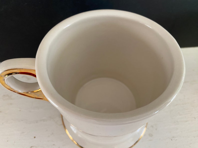 May include: White ceramic shaving mug with a gold rim and handle. The mug has a cylindrical shape and a wide, shallow base.