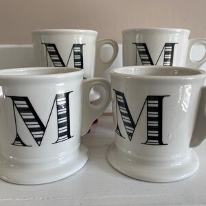 Puede incluir: Cuatro tazas de cer&aacute;mica blanca con un dise&ntilde;o de la letra "M" en negro. La letra "M" est&aacute; decorada con rayas blancas y negras. Cada taza tiene un asa y una base ancha.
