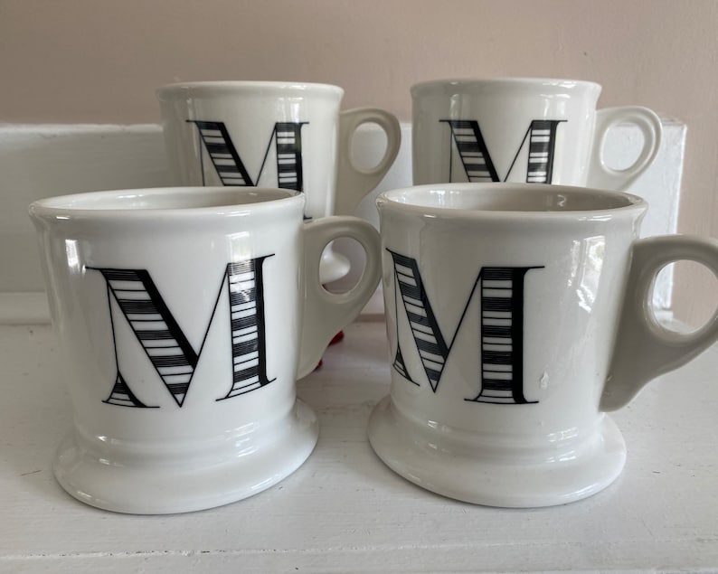 Puede incluir: Cuatro tazas de cer&aacute;mica blanca con un dise&ntilde;o de la letra "M" en negro. La letra "M" est&aacute; decorada con rayas blancas y negras. Cada taza tiene un asa y una base ancha.