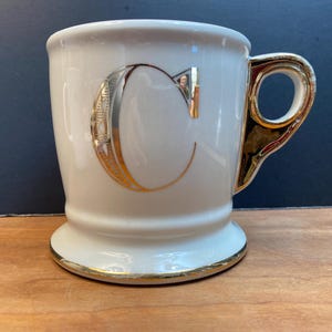 May include: White ceramic mug with a gold rim and a gold letter "C" on the front.