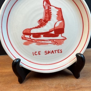 May include: A white ceramic plate with a red border and a red illustration of an ice skate. The words "ICE SKATES" are printed in red below the image. The plate is displayed on a black stand.