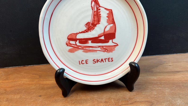 May include: A white ceramic plate with a red border and a red illustration of an ice skate. The words "ICE SKATES" are printed in red below the image. The plate is displayed on a black stand.