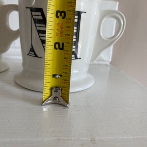 Puede incluir: Taza de cer&aacute;mica blanca con un dise&ntilde;o de la letra "N" en negro. La taza mide aproximadamente 9 cm de alto, con un asa y una base. Una cinta m&eacute;trica amarilla se coloca junto a la taza.