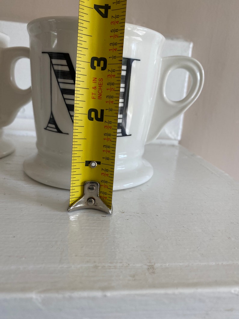 Puede incluir: Taza de cer&aacute;mica blanca con un dise&ntilde;o de la letra "N" en negro. La taza mide aproximadamente 9 cm de alto, con un asa y una base. Una cinta m&eacute;trica amarilla se coloca junto a la taza.