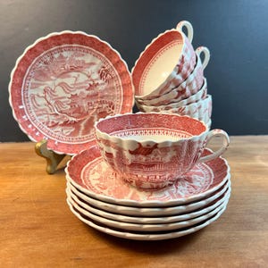 May include: A set of red and white ceramic teacups and saucers featuring a detailed transferware pattern of pagodas, trees, and bridges. The scalloped-edge saucers are stacked, and the teacups have matching red patterns with white interiors.