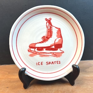 May include: A white ceramic plate with a red illustration of an ice skate. The plate has a red rim and the words "ICE SKATES" in red. The plate is displayed on a black stand.