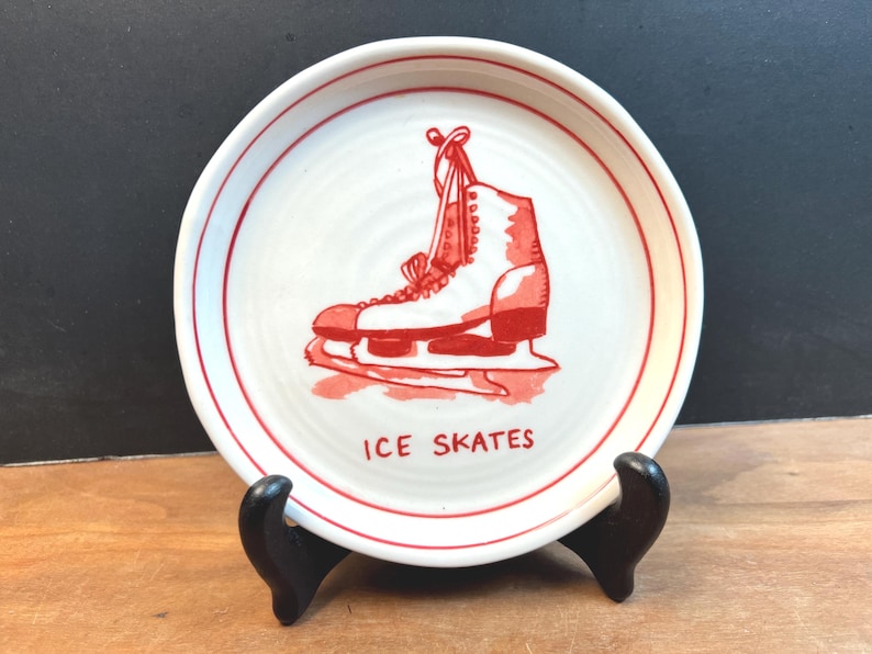 May include: A white ceramic plate with a red illustration of an ice skate. The plate has a red rim and the words "ICE SKATES" in red. The plate is displayed on a black stand.
