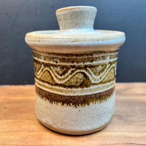 May include: A ceramic butter dish with a lid. The dish is beige with brown and olive green geometric patterns around the middle. The lid has a small handle. The dish sits on a wooden surface.