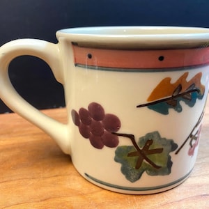 May include: Cream ceramic mug with a curved handle, decorated with purple grapes and green leaves. The rim has a red band with black dots. A blue line accents the base. The mug is suitable for hot drinks.