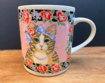 May include: A white ceramic mug with a pink background and a colorful floral design. A tabby cat with a blue bow is featured in the center of the mug.