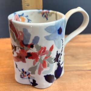 May include: White ceramic mug with a floral design in red, blue, and pink. The mug has a curved handle and a unique, slightly scalloped base. The flowers are painted in a watercolor style, adding a delicate touch to this kitchenware item.