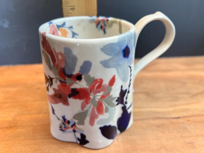 May include: White ceramic mug with a floral design in red, blue, and pink. The mug has a curved handle and a unique, slightly scalloped base. The flowers are painted in a watercolor style, adding a delicate touch to this kitchenware item.