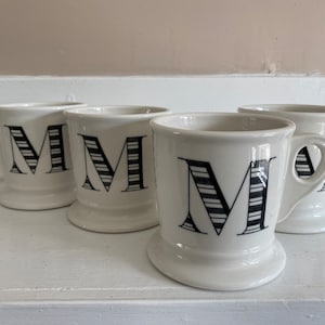Puede incluir: Cuatro tazas de cer&aacute;mica blanca con una letra "M" a rayas blancas y negras. Cada taza tiene un asa y una base redondeada. Las tazas est&aacute;n dispuestas sobre una superficie blanca.