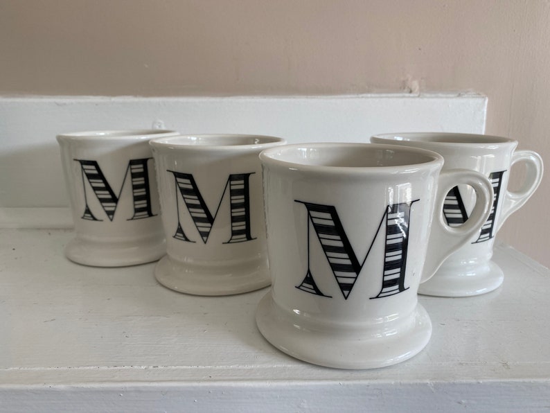 Puede incluir: Cuatro tazas de cer&aacute;mica blanca con una letra "M" a rayas blancas y negras. Cada taza tiene un asa y una base redondeada. Las tazas est&aacute;n dispuestas sobre una superficie blanca.