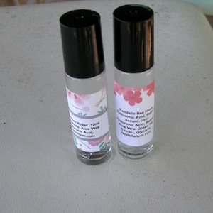 May include: Two clear glass roller bottles with black caps and labels. The labels are white with pink flowers and text that reads "Randalia Bee Hives Hyaluronic Acid, Retinol Serum - 10ml Hyaluronic Acid, Retinol, Aloe Vera, Green Tea Extract, Glycerin, randaliafarm.com".